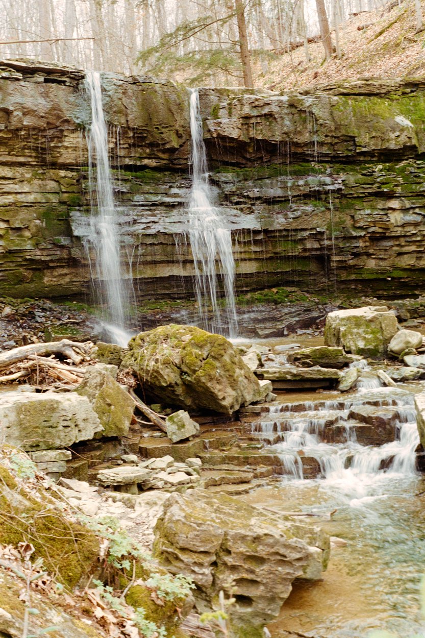 A serene forest waterfall scene featuring water cascading down a layered, moss-covered rock cliff face from two main falls. The water tumbles into a rocky pool below, flowing over tiered stone ledges and boulders into a stream. Surrounding the falls are mossy rocks, fallen branches, and a backdrop of bare trees, suggesting late fall or early spring in a quiet woodland setting.